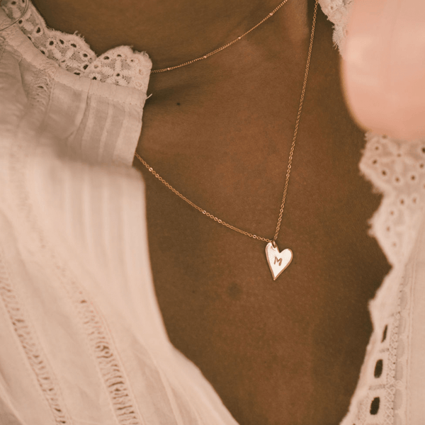 A close-up of a woman in a white lace top wearing a dainty gold choker layered with a delicate gold pendant chain necklace with a flat polished heart charm, engraved with the letter 'M'.