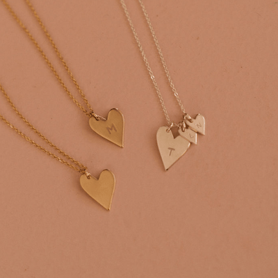 Three delicate pendant chain necklaces, two gold and one silver, laying on a solid colored blush pink surface. The two gold each have one flat gold heart charm, with one being engraved with the letter 'M'. The silver has a larger silver heart charm with the letter 'T', and two smaller silver heart charms that each have a letter engraved.