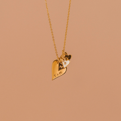 A delicate gold pendant chain necklace with a large flat heart charm and multiple smaller flat heart charms. The large charm is engraved with 'T + M' and the smaller hearts each have a single initial.