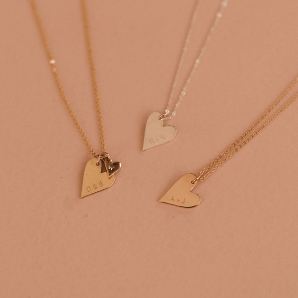 Three delicate pendant chain necklaces in gold, silver, and rose gold, each with a large engraved flat heart charm. The gold necklace also has multiple smaller heart charms, each engraved with an initial. All of the necklaces are laying on a blush colored solid surface.