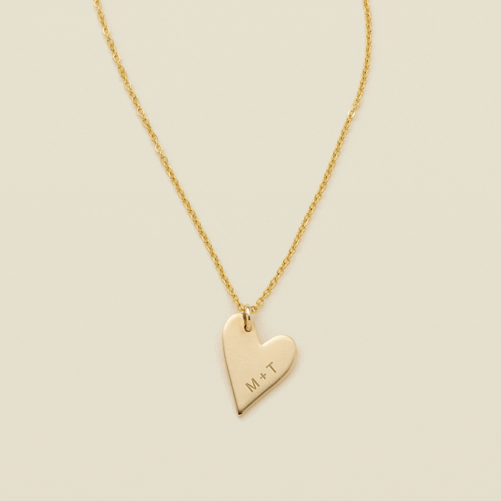 A gift of a delicate gold pendant chain necklace, starting with a large flat heart charm engraved with 'M + T', then adding multiple smaller flat heart charms. Each miniature heart charm is engraved with a single initial.