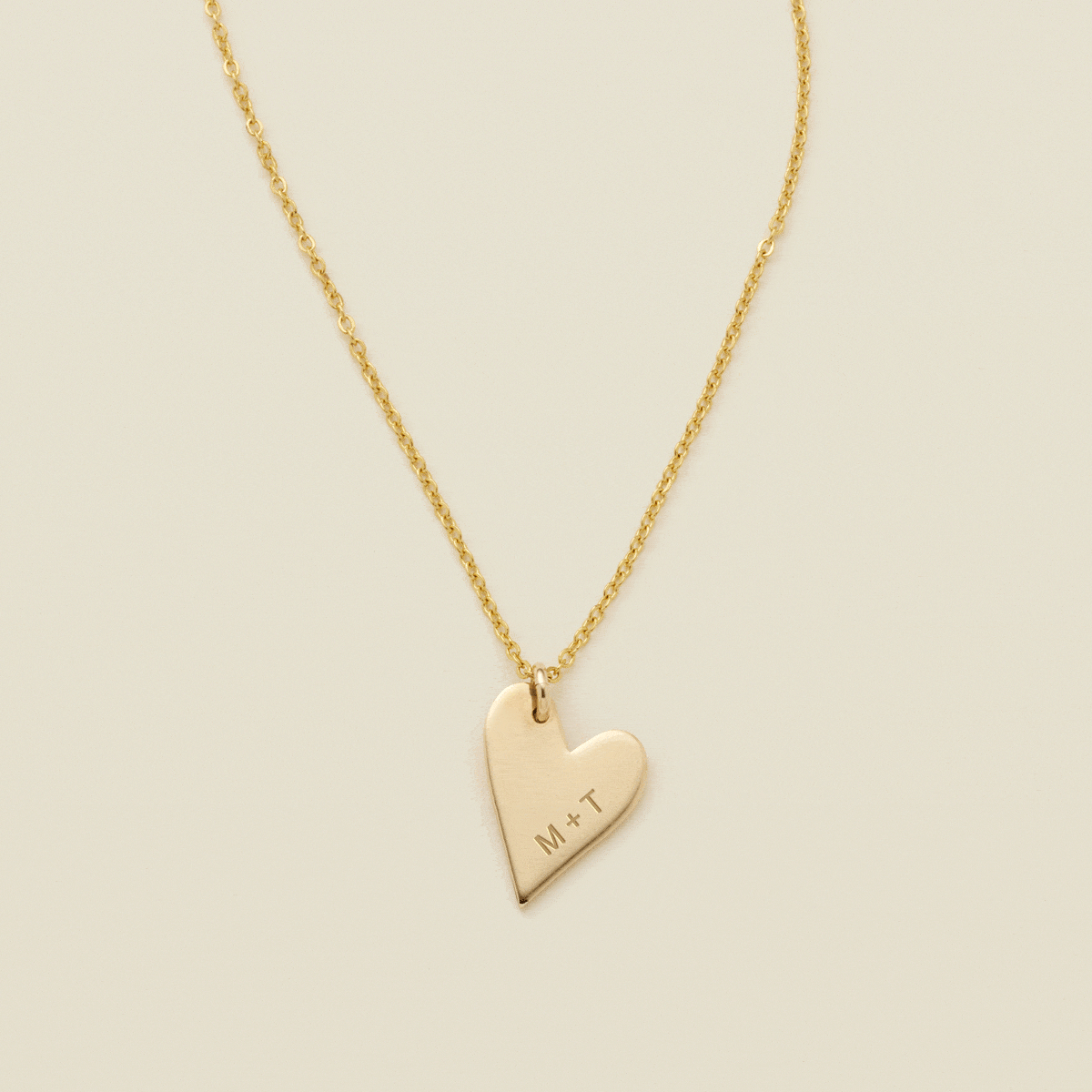 A gift of a delicate gold pendant chain necklace, starting with a large flat heart charm engraved with 'M + T', then adding multiple smaller flat heart charms. Each miniature heart charm is engraved with a single initial.
