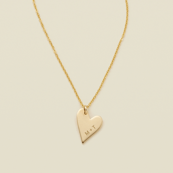 A gift of a delicate gold pendant chain necklace, starting with a large flat heart charm engraved with 'M + T', then adding multiple smaller flat heart charms. Each miniature heart charm is engraved with a single initial.