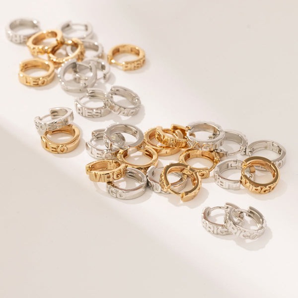 Multiple gold and silver huggie hoop earrings scattered on a solid white surface. Each earring has a different zodiac sign name in raised letters across the front.