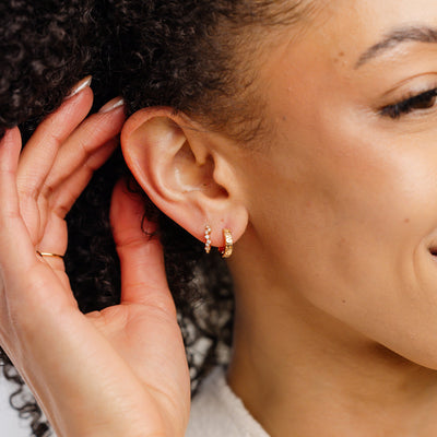 A close-up of a person's ear wearing two gold hoop earrings, one with clear round gemstones in a curved pattern and the other with the word 'TAURUS' in raised letters across the front.