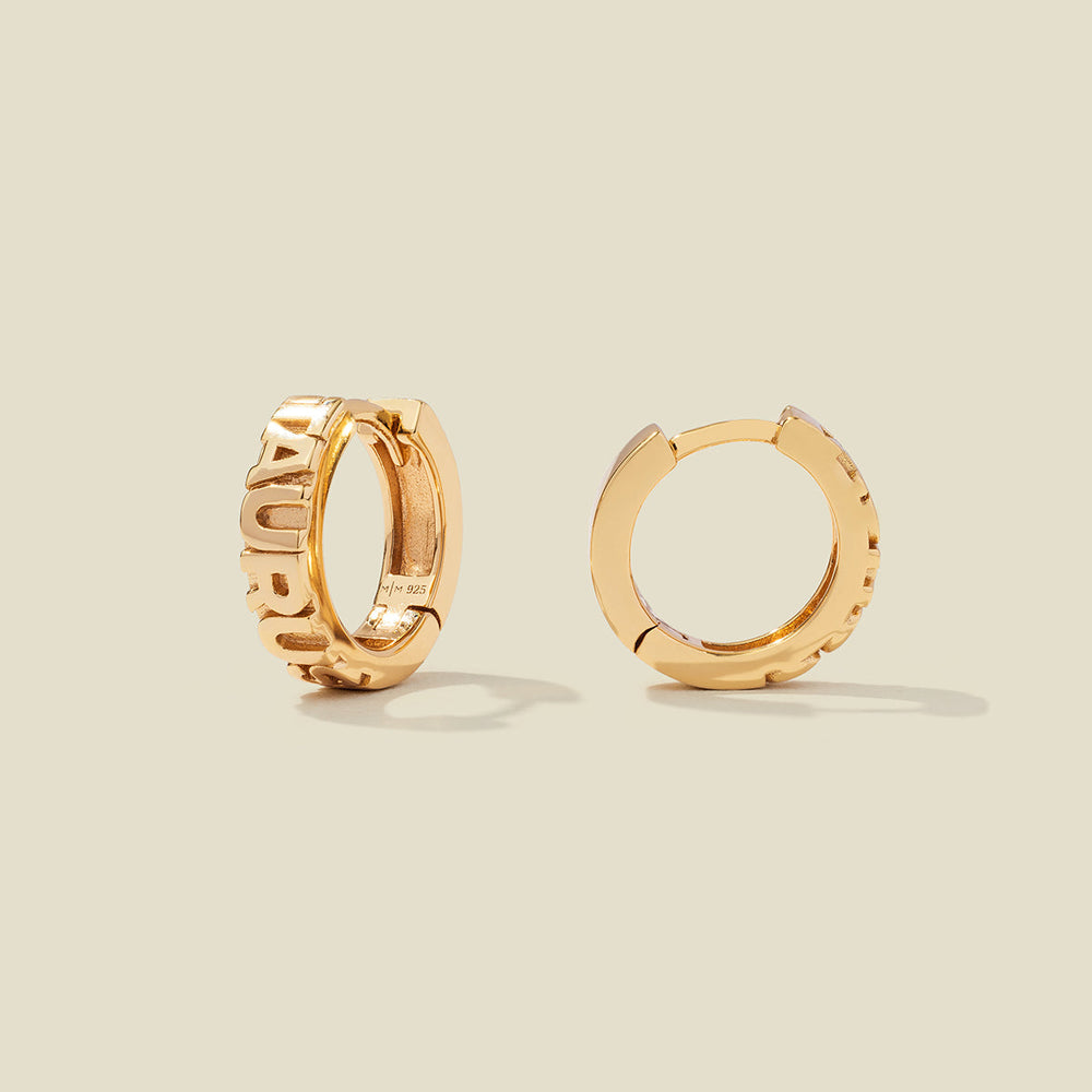 A pair of gold huggie hoop earrings on a light beige background. One earring is shown from the side, displaying a polished outer surface, while the other is shown from the front, showcasing the word 'TAURUS' in raised letters.