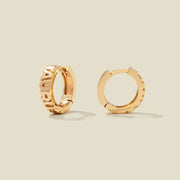 A pair of gold huggie hoop earrings on a light beige background. One earring is shown from the side, displaying a polished outer surface, while the other is shown from the front, showcasing the word 'TAURUS' in raised letters.