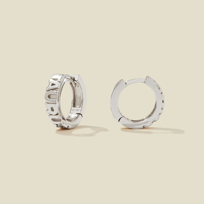 A pair of silver huggie hoop earrings on a light beige background. One earring is shown from the side, displaying a polished outer surface, while the other is shown from the front, showcasing the word 'TAURUS' in raised letters.