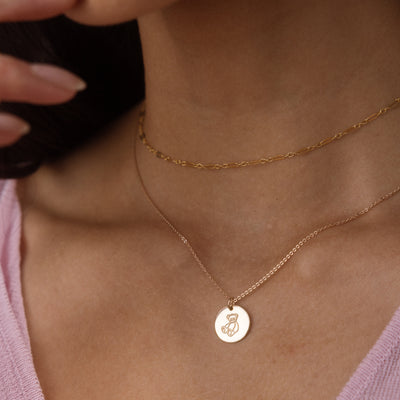 A close-up of a person's neck and chest wearing two delicate gold necklaces: a thin chain necklace with a round engraved teddy bear disc pendant and a slightly thicker chain necklace above it.