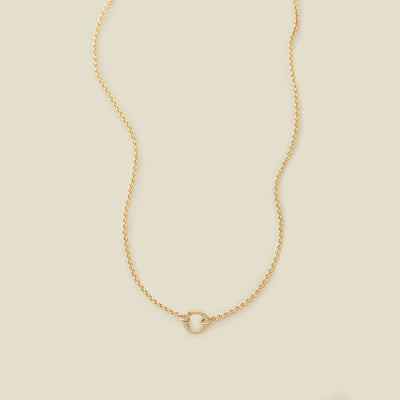 A gold circular link chain necklace with a round ring in the center, shown on a solid light beige background.