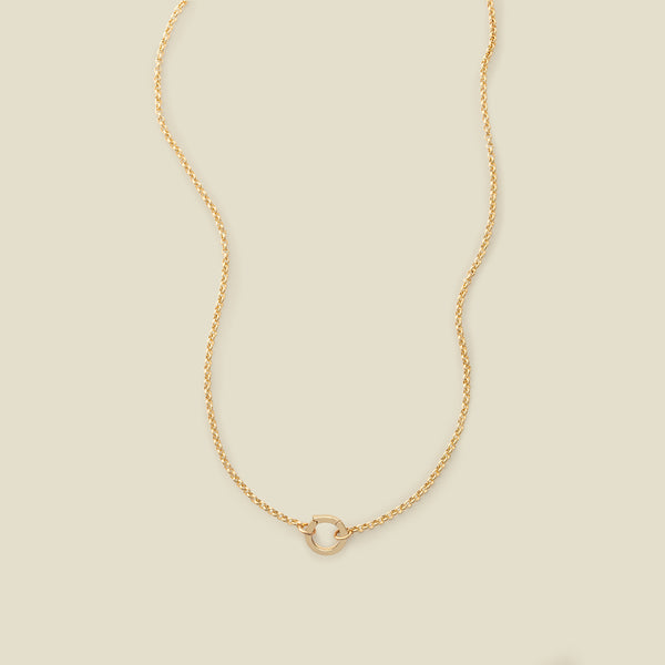 A gold circular link chain necklace with a round ring in the center, shown on a solid light beige background.
