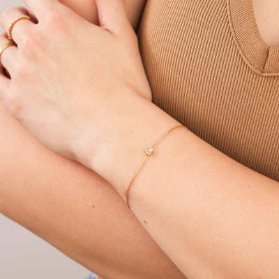 A close-up of a person wearing a brown ribbed top with their arms crossed across their chest, showcasing a delicate gold chain bracelet with baguette and pear cut clear prong set gemstones.