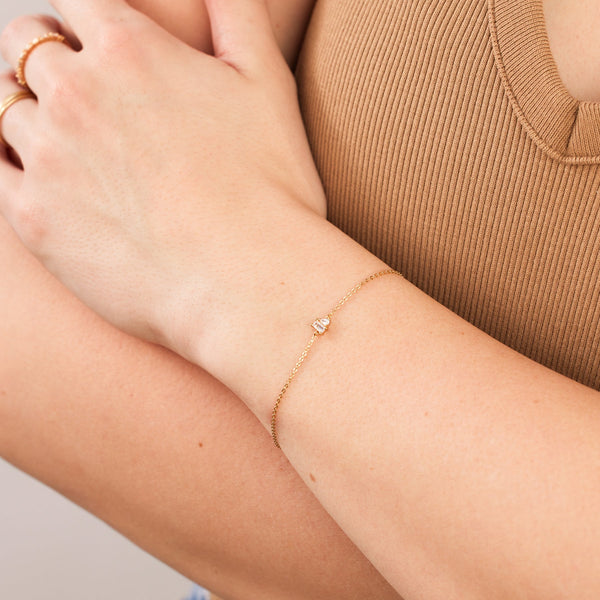 A close-up of a person wearing a brown ribbed top with their arms crossed across their chest, showcasing a delicate gold chain bracelet with baguette and pear cut clear prong set gemstones.
