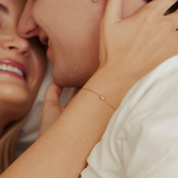 A close-up of a person holding another persons face, showcasing a gold delicate chain bracelet with a clear baguette and clear pear prong set gemstones side-by-side.