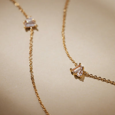 A close-up of two delicate gold chain bracelets, each with a clear baguette and clear pear cut prong set gemstone side-by-side, shown on a light beige background with shadows.