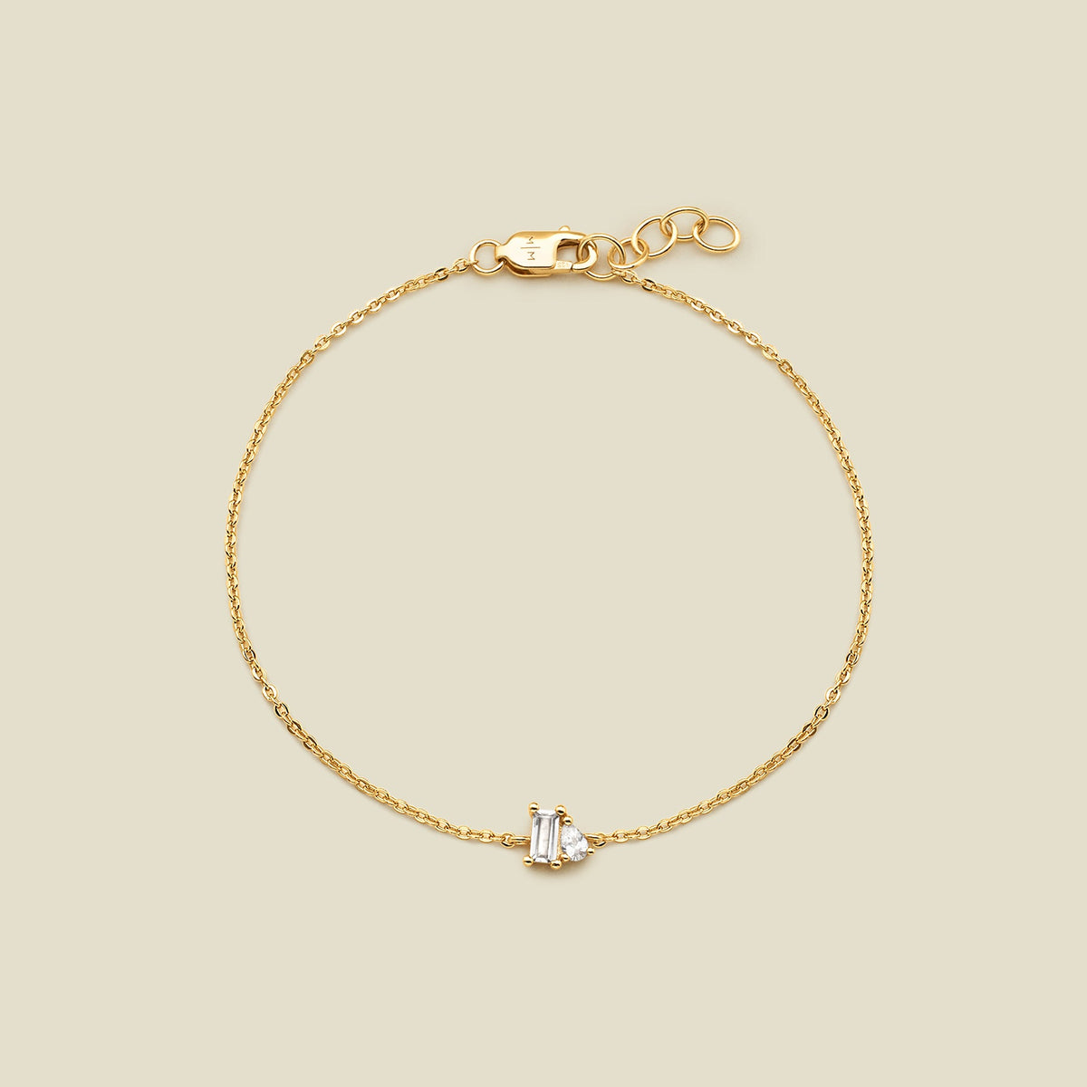 A delicate gold chain bracelet with a small central clear baguette-cut gemstone and pear-cut clear gemstone in a prong setting, displayed on a solid light beige background.