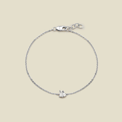 A delicate silver chain bracelet with a small central clear baguette-cut gemstone and pear-cut clear gemstone in a prong setting, displayed on a solid light beige background.
