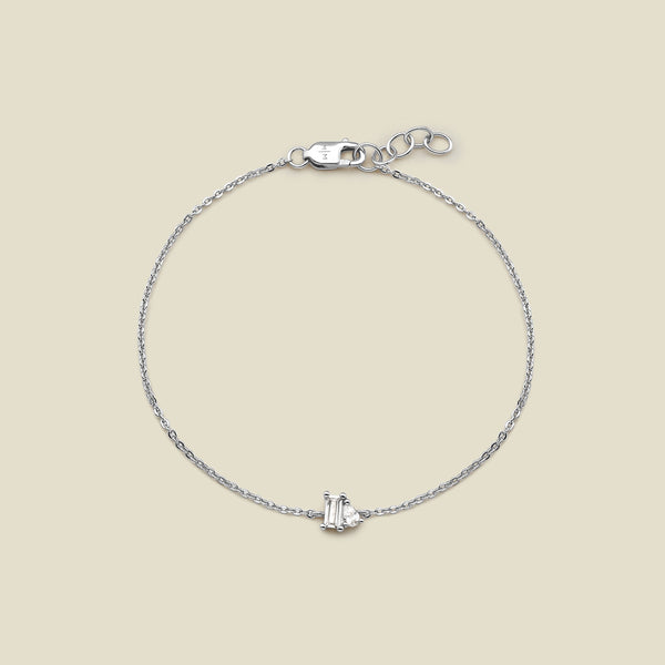 A delicate silver chain bracelet with a small central clear baguette-cut gemstone and pear-cut clear gemstone in a prong setting, displayed on a solid light beige background.