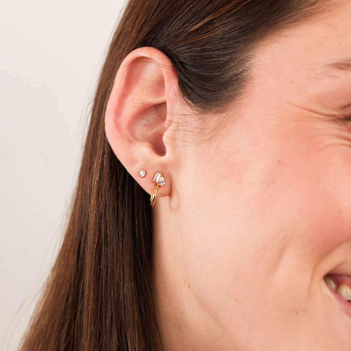 A close-up of a person's ear wearing a gold hoop with pear and baguette cut clear gemstones, and a single prong set round clear gemstone stud.