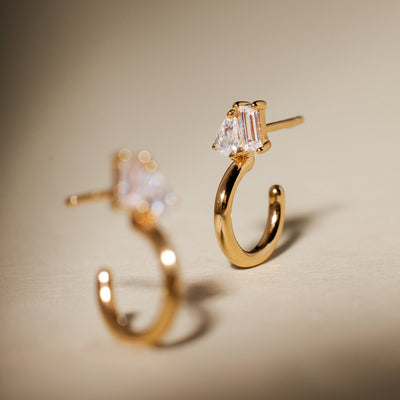 A gold hoop earring featuring pear and baguette cut clear gemstones in prong settings side-by-side, with the second hoop blurred in the foreground.