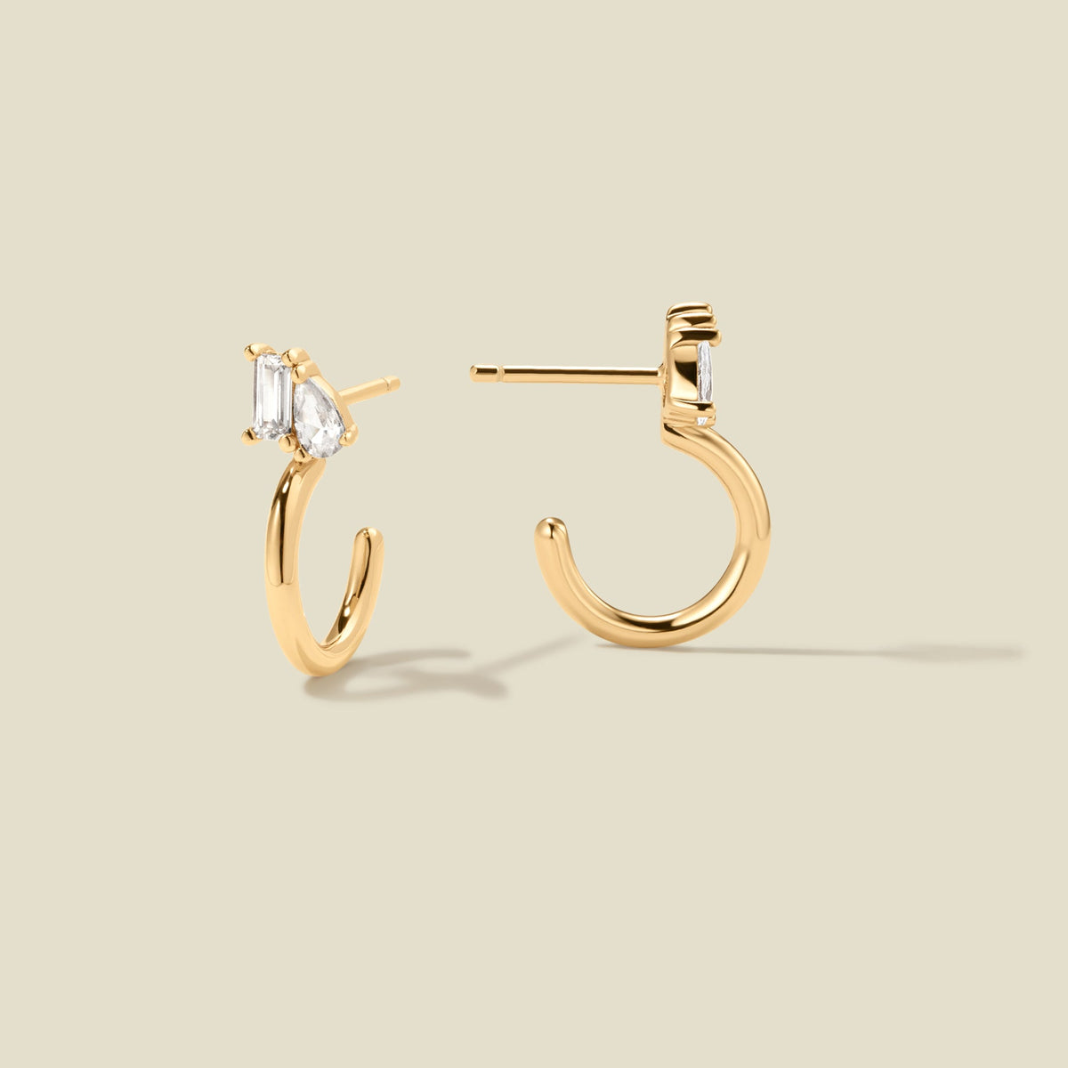 A pair of gold hoop earrings with clear pear and baguette-cut stones, shown from two angles on a solid beige background.
