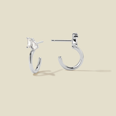 A pair of silver hoop earrings with clear pear and baguette-cut stones, shown from two angles on a beige background.