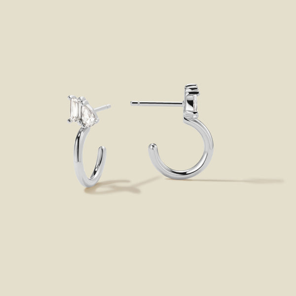 A pair of silver hoop earrings with clear pear and baguette-cut stones, shown from two angles on a beige background.