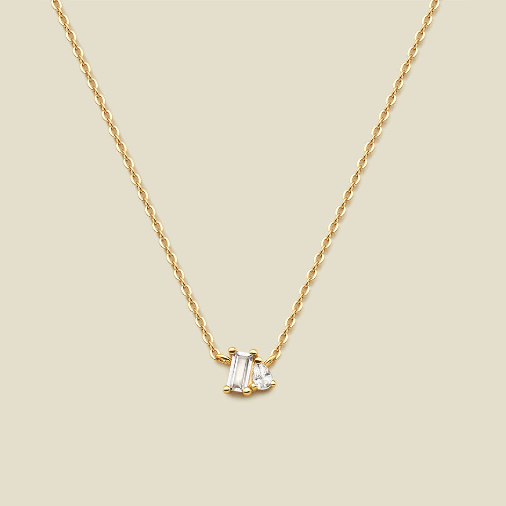A delicate gold chain necklace featuring a small pendant with a baguette-cut clear gemstone and a small pear cut clear gemstone, set against a solid beige background.