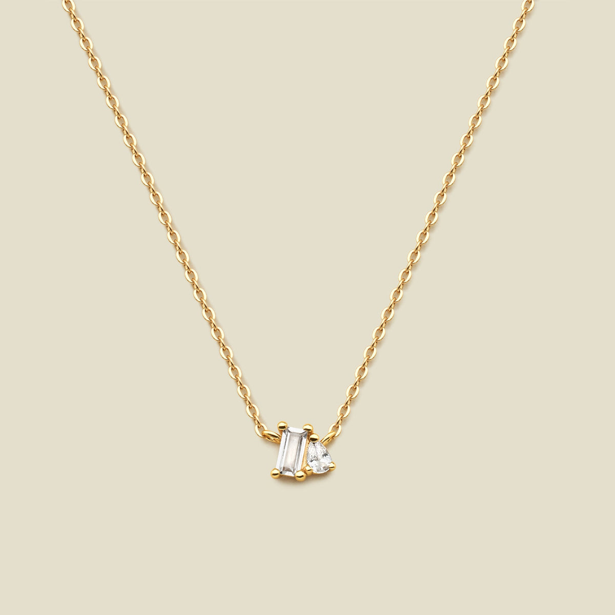 A delicate gold chain necklace featuring a small pendant with a baguette-cut clear gemstone and a small pear cut clear gemstone, set against a solid beige background.