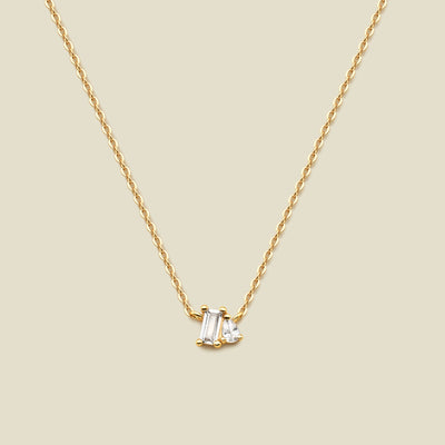 A delicate gold chain necklace featuring a small pendant with a baguette-cut clear gemstone and a small pear cut clear gemstone, set against a solid beige background.