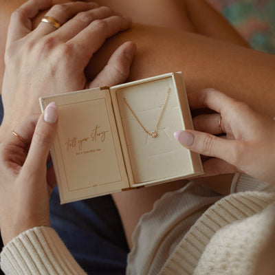 A close-up of a person holding a jewelry box open with the words "tell your story" and "it's a beautiful one" on the inside. The box holds a delicate gold chain necklace with clear baguette and pear cut gemstones side-by-side in the center.