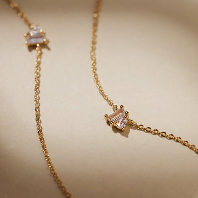 A close-up of two delicate gold chain necklaces, each with clear baguette and pear cut gemstones side-by-side in prong settings in the center, laying on a solid light beige surface.