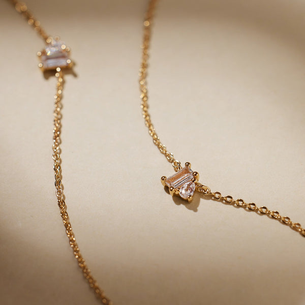 A close-up of two delicate gold chain necklaces, each with clear baguette and pear cut gemstones side-by-side in prong settings in the center, laying on a solid light beige surface.