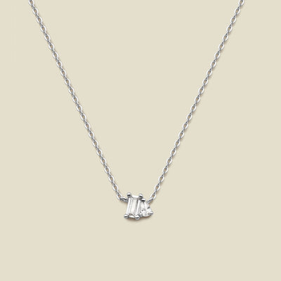A delicate silver chain necklace featuring a small pendant with a baguette-cut clear gemstone and a small pear cut clear gemstone, set against a solid beige background.