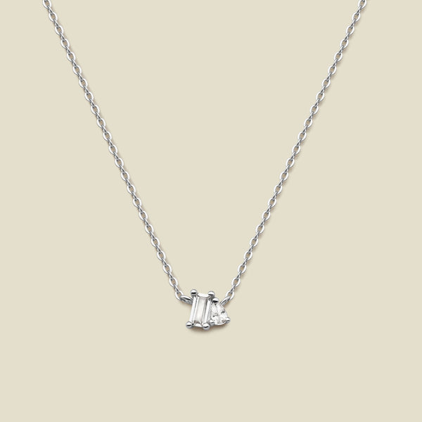 A delicate silver chain necklace featuring a small pendant with a baguette-cut clear gemstone and a small pear cut clear gemstone, set against a solid beige background.