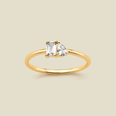 A gold ring featuring a rectangular clear gemstone and a pear-cut clear gemstone, shown on a solid light beige background. 