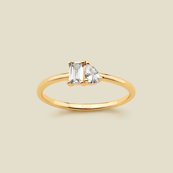 A gold ring featuring a rectangular clear gemstone and a pear-cut clear gemstone, shown on a solid light beige background. 