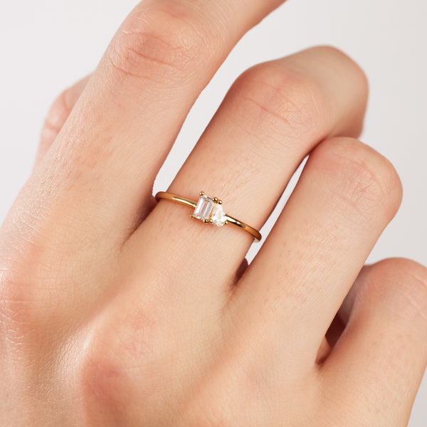A close-up of a person's hand wearing a thin gold band ring with a baguette-cut and pear-cut clear gemstone in prong settings side-by-side, set against a solid white background.
