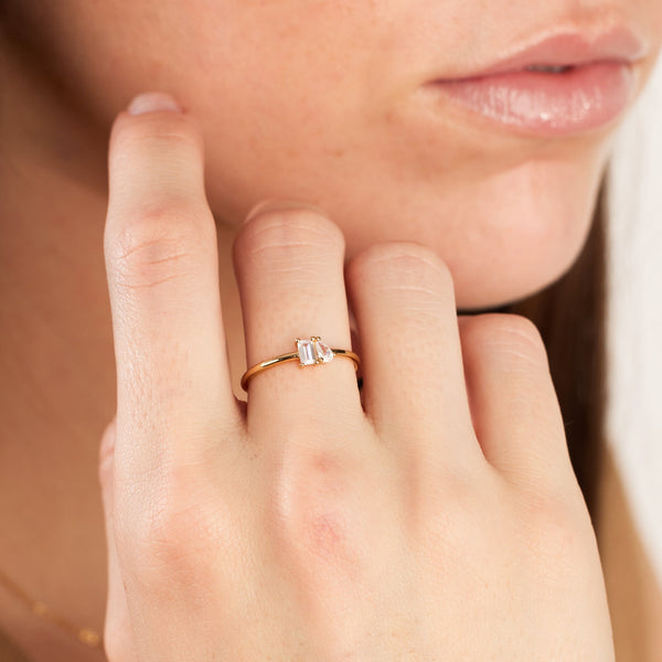 A close-up of a person resting their hand on their chin, showing a thin band gold ring with prong set baguette and pear clear gemstones.