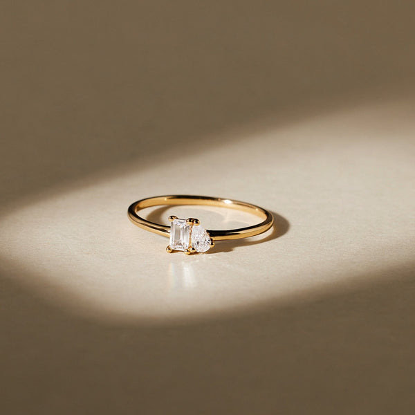 A thin gold band ring featuring a baguette-cut and pear-cut clear gemstone side-by-side in prong settings, shown on a light beige surface with shadows.