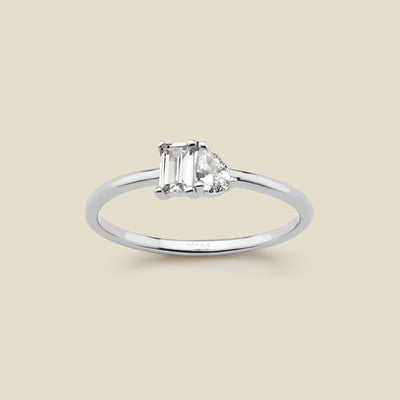 A silver ring featuring a rectangular clear gemstone and a pear-cut clear gemstone, shown on a solid light beige background. 