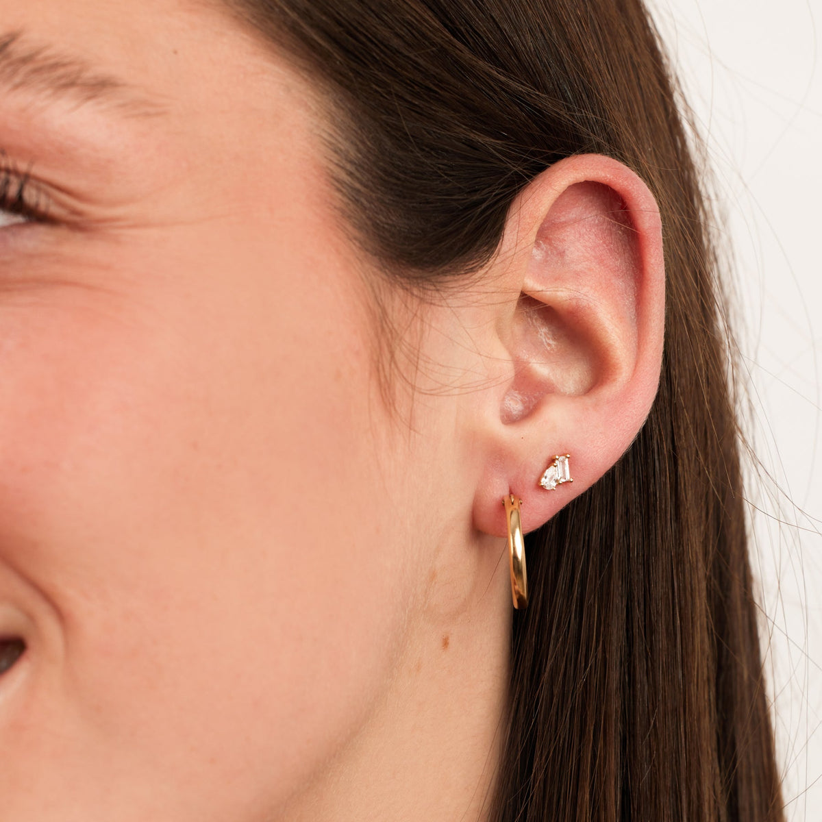 A close-up of a person's ear wearing a polished gold hoop and a gold prong stud with a pear-cut and baguette-cut clear gemstone side by side, set against a solid white background.