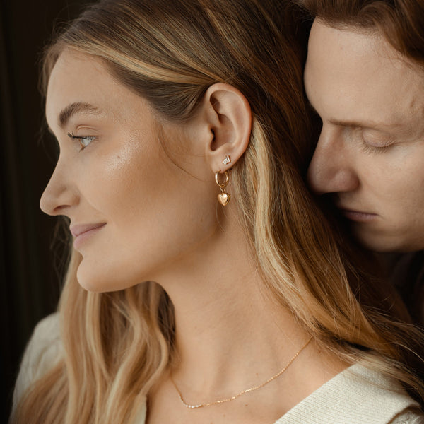 A close-up of a woman with a man hugging her from behind, showcasing a gold hoop earring with a polished gold heart dangling from it, and a gold stud with clear gemstones.