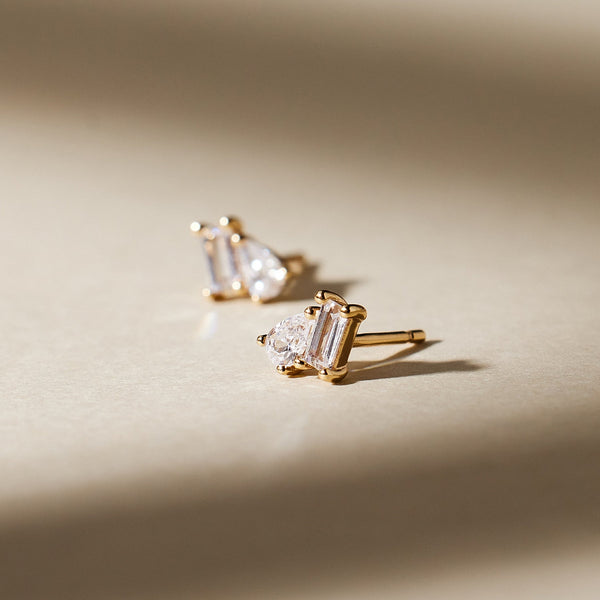 A prong set gold stud earring featuring a pear-cut and baguette-cut clear gemstone set side by side, shown on a light beige surface with moody shadows.