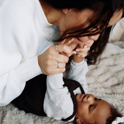 A close-up of a mother playing with her child who is laying flat on the ground, grabbing the child's hands near her face and smiling.