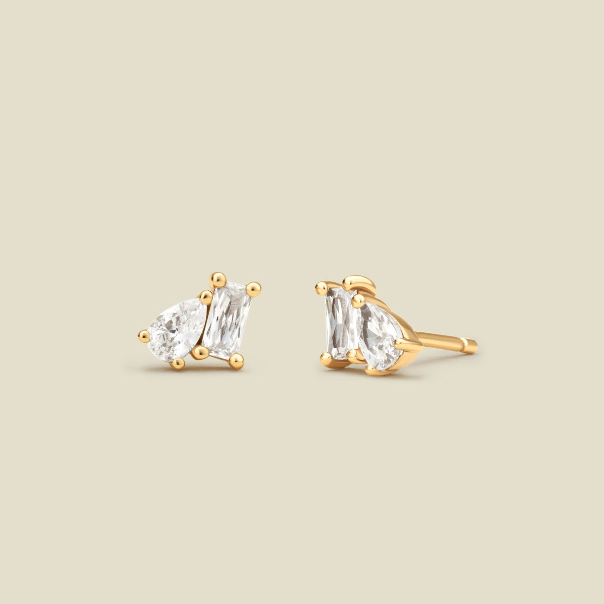 A pair of gold stud earrings with baguette and pear-cut clear gemstones in prong settings, shown on a solid light beige background.