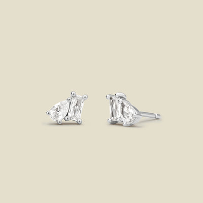 A pair of silver stud earrings with baguette and pear-cut clear gemstones in prong settings, shown on a solid light beige background.