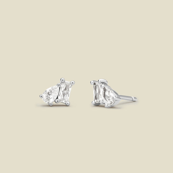 A pair of silver stud earrings with baguette and pear-cut clear gemstones in prong settings, shown on a solid light beige background.