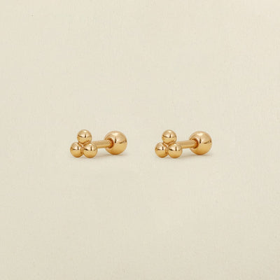 A pair of gold stud earrings with three spherical beads, displayed on a light beige textured background.