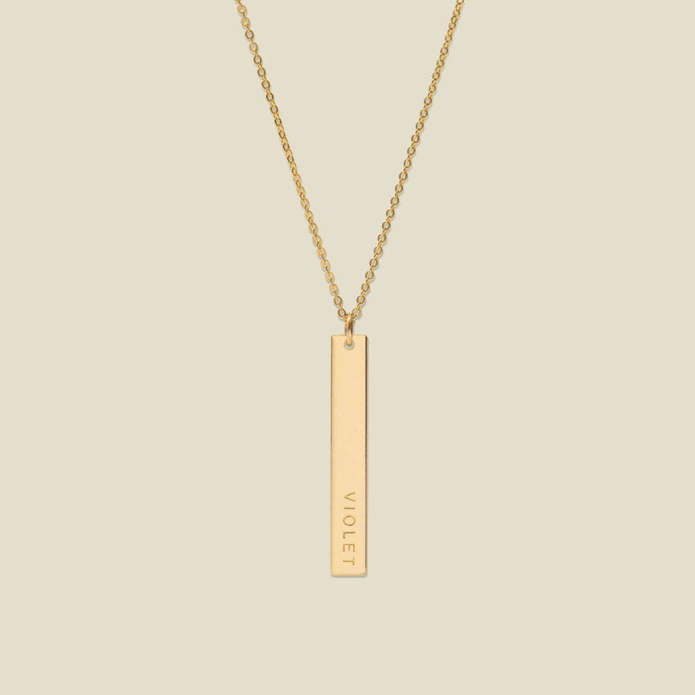 A gold pendant necklace with a vertical bar engraved with the name 'VIOLET' on a light beige background.